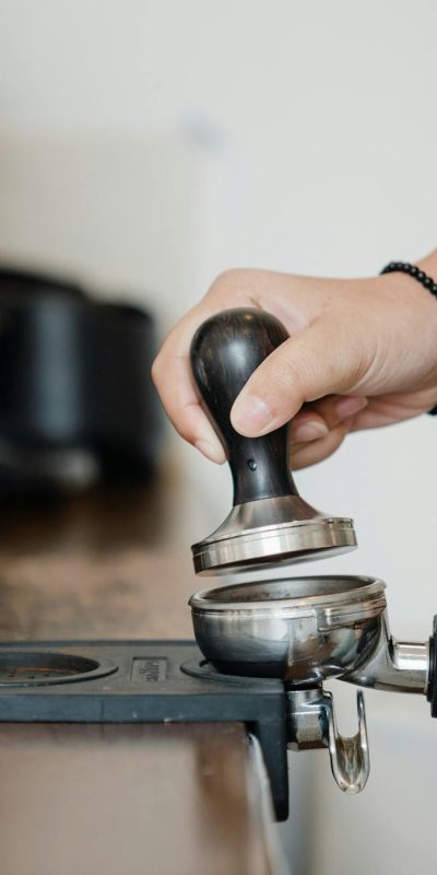 Crop anonymous barista pressing aromatic coffee beans into filter block by using tamper while standing in light kitchen nearby professional brewing machine