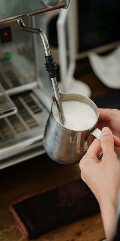 Crop cafe worker using coffeemaker to whip milk in pitcher