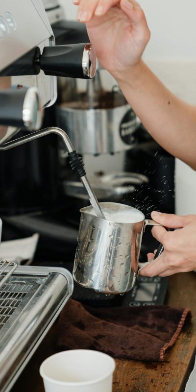 Crop faceless young female barista in apron steaming milk in steel pot using modern cappuccino machine