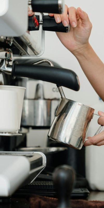 Crop faceless young female barista in apron steaming milk at modern coffee machine while preparing delicious latte